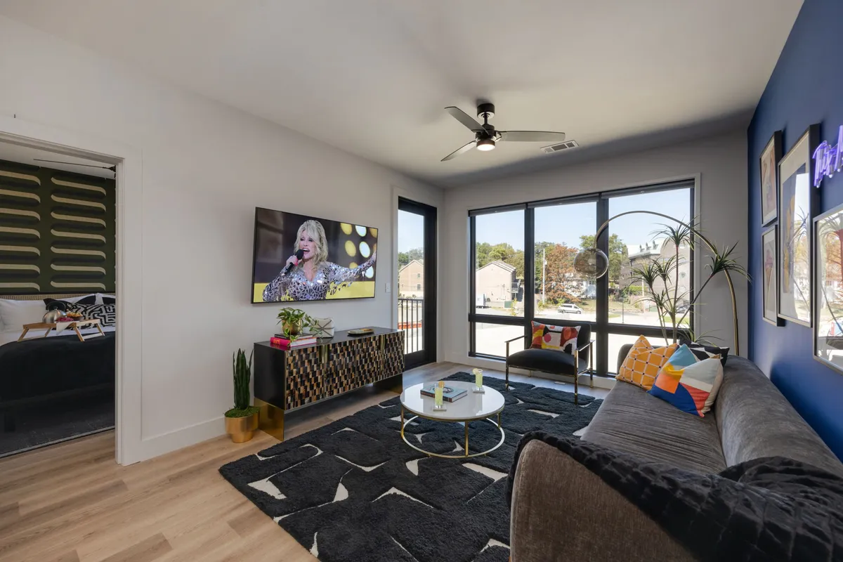 A sun-filled living room with a colorful couch facing a television displaying a lively performance. Large windows allow ample natural light to spill into the space, and vibrant decorative touches add to the cheerful ambiance. The room feels designed for both relaxation and entertainment, with plush seating and a visible Smart TV.