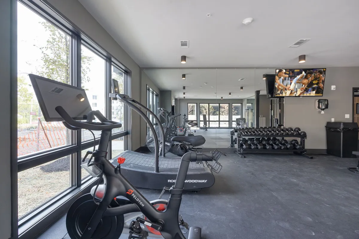 A bright, spacious fitness center interior with a Peloton bike, Woodway treadmill, dumbbell rack, large windows, and a TV screen. The natural light and state-of-the-art equipment are prominent.