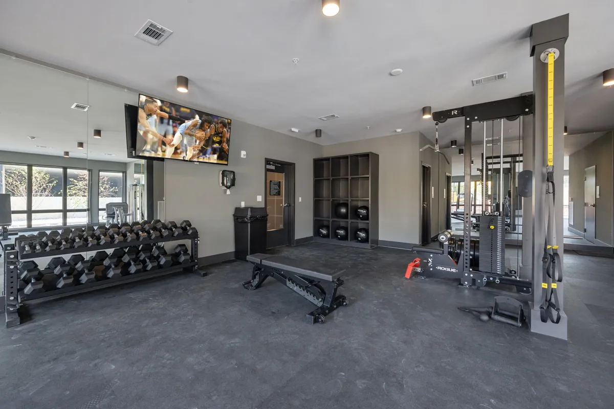 An interior view of the fitness center, showcasing a range of modern exercise equipment including dumbbells, a weight bench, exercise equipment, and a television screen. The space is designed to be motivating and energetic, ideal for personal workouts and community fitness events.