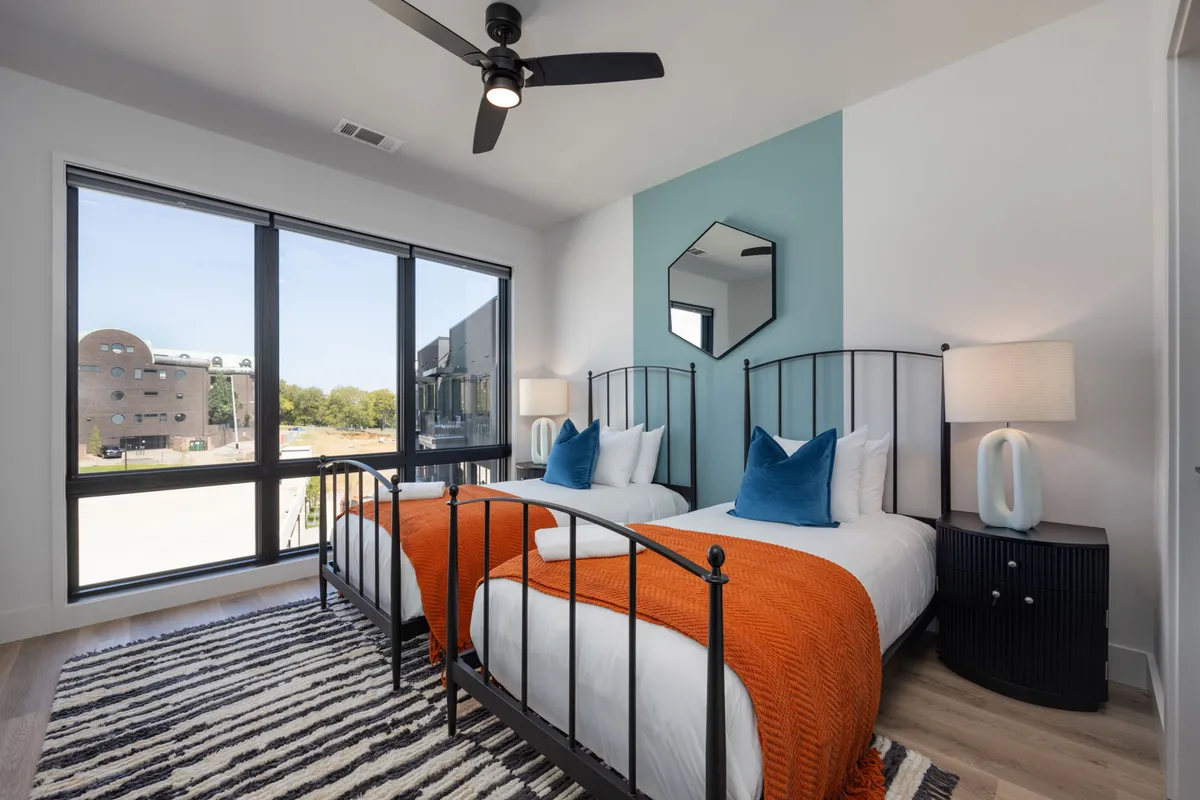 A comfortable and modern bedroom with two twin beds, a geometric mirror, striped rug, and decorative lamps. The space is bright and designed for both rest and togetherness, making it ideal for families or friends.