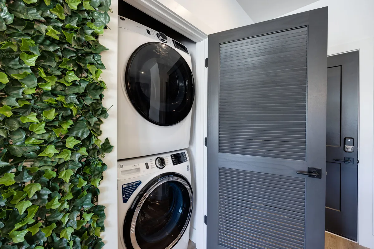 A laundry area featuring a stacked washer and dryer, accented by a green ivy wall and a modern door. The space is practical, well-organized, and visually refreshing, emphasizing day-to-day convenience.