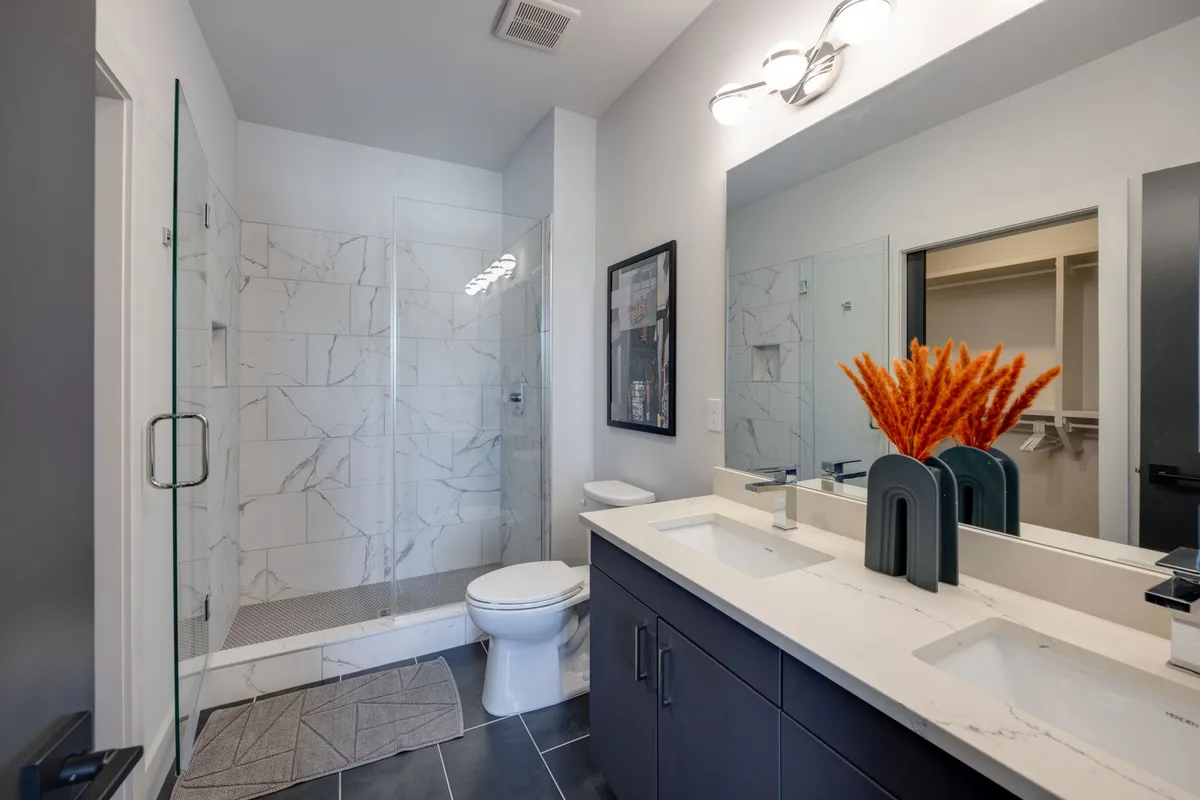 A fresh bathroom with a double sink vanity, glass shower enclosure, and a decorative vase with orange flowers. The finishes are modern, and the design evokes a spa-like experience, ideal for self-care and rejuvenation.