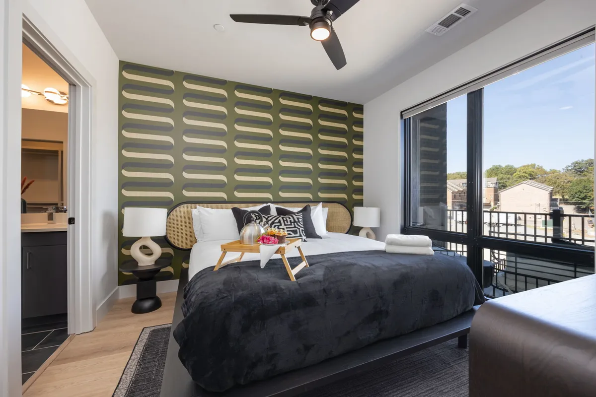 A stylish bedroom featuring a large bed with dark bedding, decorative pillows, unique wallpaper, nightstands with lamps, and a view through expansive windows. The natural light gives the room a serene, inviting atmosphere, ideal for relaxation.